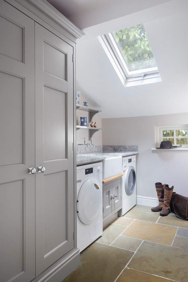 Inspiration för små klassiska linjära grått tvättstugor enbart för tvätt, med en rustik diskho, granitbänkskiva, en tvättmaskin och torktumlare bredvid varandra, luckor med infälld panel, grå skåp, grå väggar och travertin golv