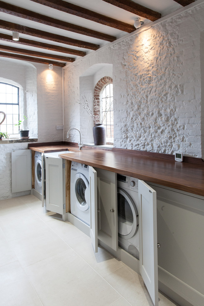 Landhaus Waschküche mit Landhausspüle, Schrankfronten im Shaker-Stil, grauen Schränken, Arbeitsplatte aus Holz, weißer Wandfarbe, Waschmaschine und Trockner versteckt und brauner Arbeitsplatte in Hampshire