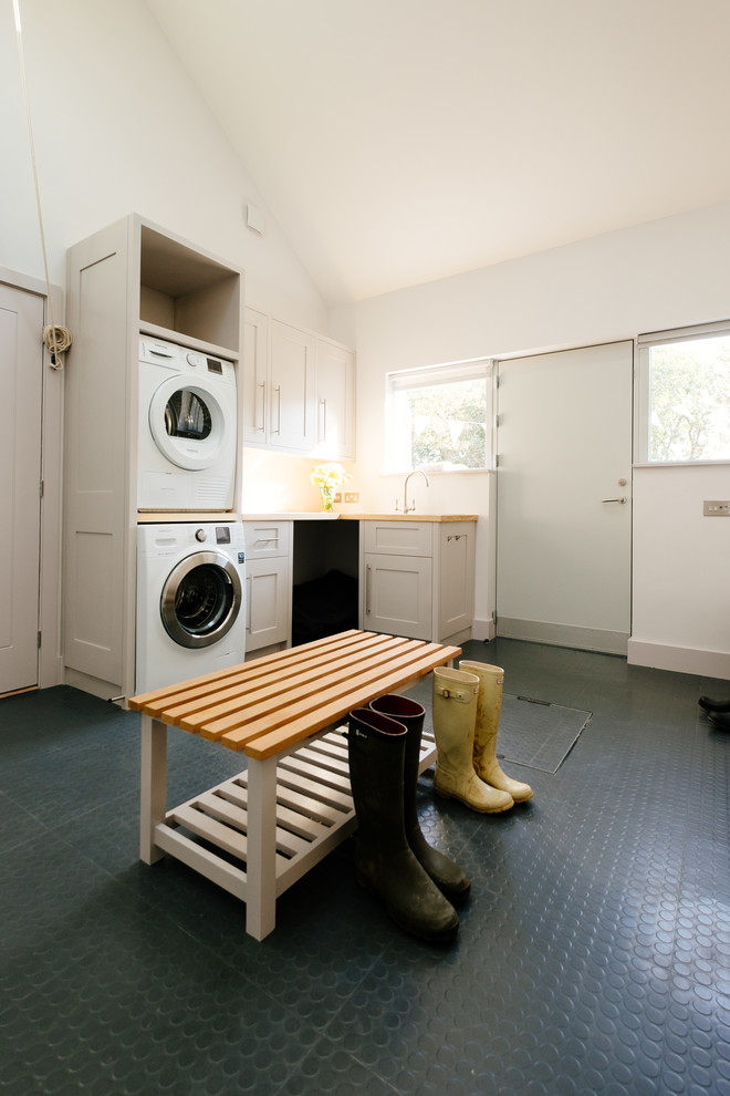 Klassischer Hauswirtschaftsraum in L-Form mit Schrankfronten im Shaker-Stil, weißen Schränken, Arbeitsplatte aus Holz, weißer Wandfarbe, Waschmaschine und Trockner gestapelt und beiger Arbeitsplatte in Cornwall
