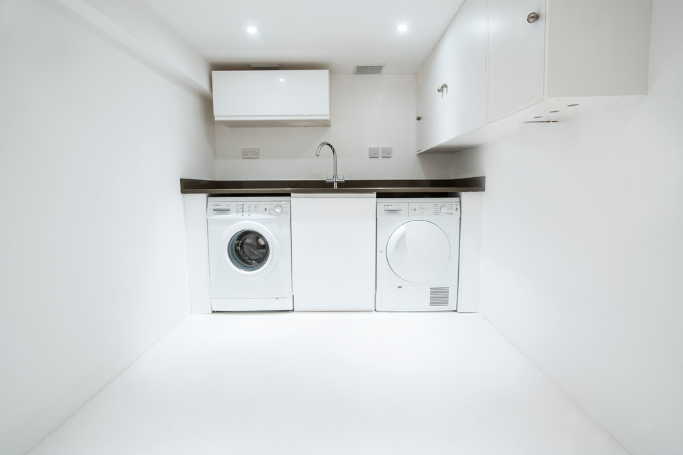Modern inredning av en mellanstor linjär tvättstuga enbart för tvätt, med en undermonterad diskho, släta luckor, vita skåp, granitbänkskiva, vita väggar, betonggolv och en tvättmaskin och torktumlare bredvid varandra