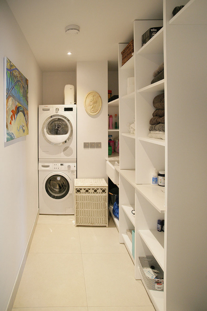 サセックスにある高級な小さなモダンスタイルのおしゃれな家事室 (I型、エプロンフロントシンク、オープンシェルフ、白いキャビネット、人工大理石カウンター、ベージュキッチンパネル、ガラス板のキッチンパネル、白い壁、セラミックタイルの床、上下配置の洗濯機・乾燥機、ベージュの床、ベージュのキッチンカウンター) の写真