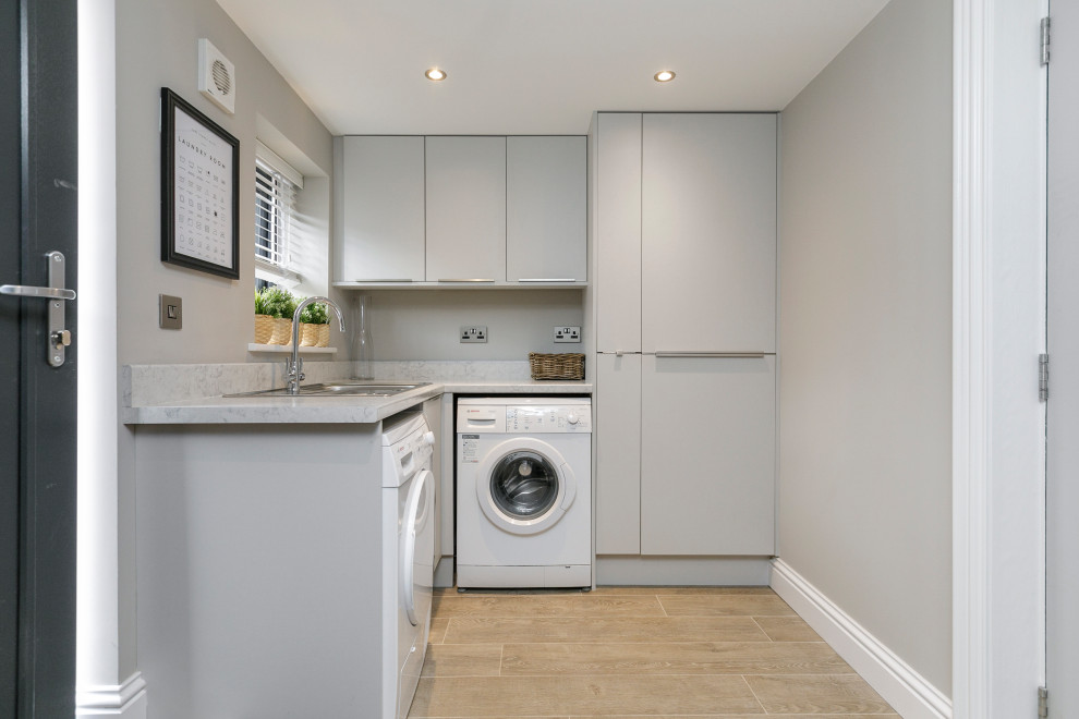 Moderne Waschküche in L-Form mit Einbauwaschbecken, flächenbündigen Schrankfronten, grauen Schränken, Quarzwerkstein-Arbeitsplatte, grauer Wandfarbe, Waschmaschine und Trockner nebeneinander, braunem Boden und grauer Arbeitsplatte in Belfast