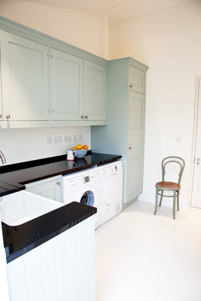 Photo of a scandi l-shaped separated utility room in London with a belfast sink, recessed-panel cabinets, blue cabinets, white walls, ceramic flooring and a side by side washer and dryer.