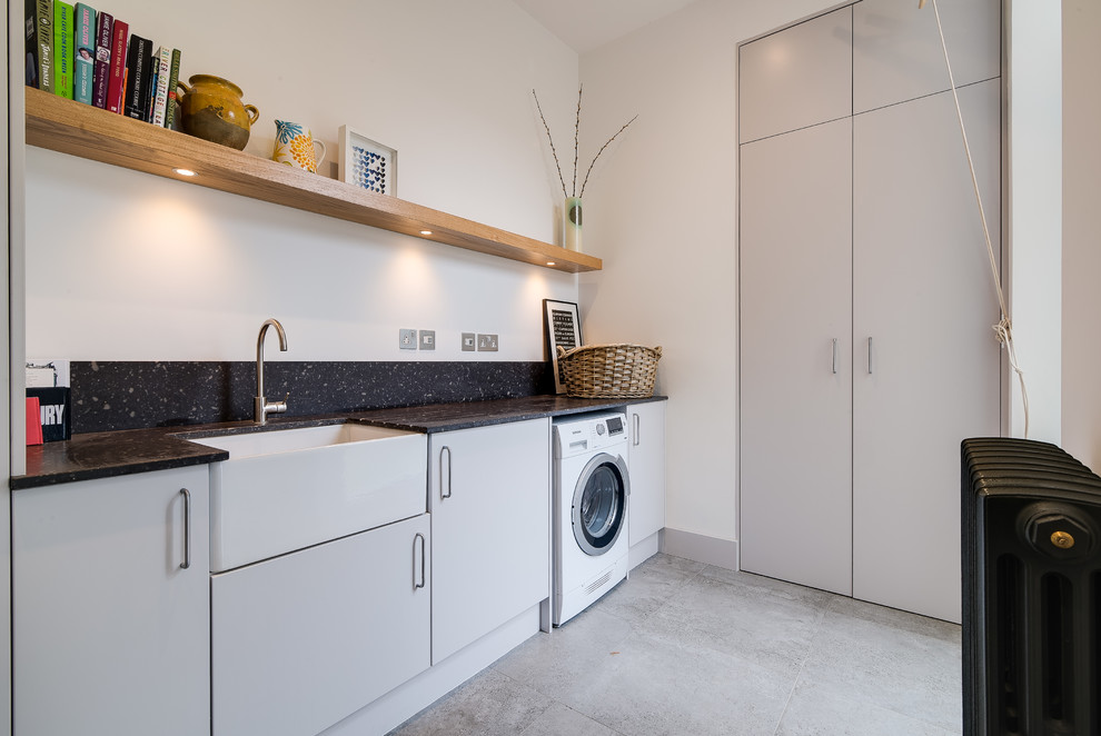 Photo of a medium sized contemporary single-wall utility room in London with a belfast sink, flat-panel cabinets, grey cabinets, granite worktops, white walls, ceramic flooring, grey floors and black worktops.