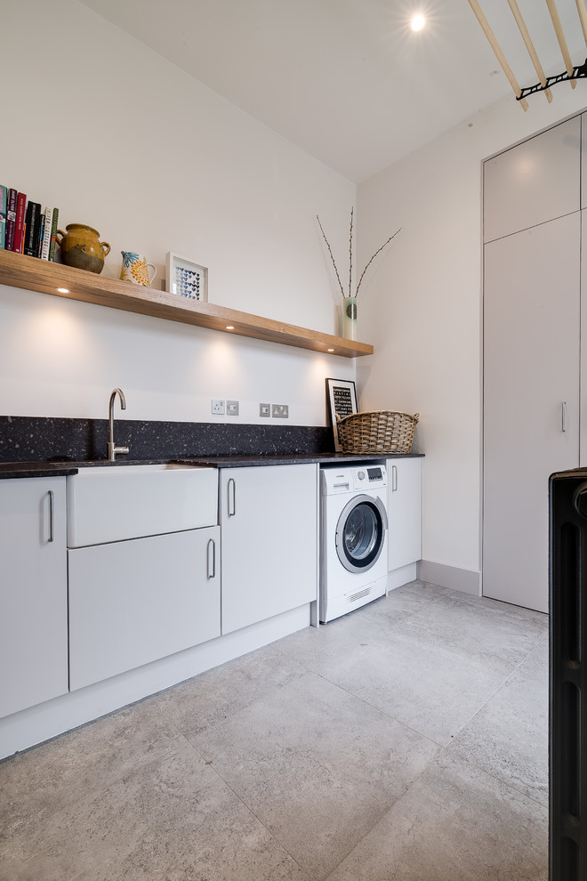 Design ideas for a medium sized contemporary single-wall laundry cupboard in London with a belfast sink, flat-panel cabinets, grey cabinets, granite worktops, white walls, ceramic flooring, grey floors and black worktops.