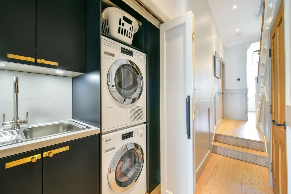 This is an example of a traditional single-wall laundry cupboard in London with a built-in sink, flat-panel cabinets, black cabinets, light hardwood flooring, a stacked washer and dryer, beige floors, white worktops and a dado rail.