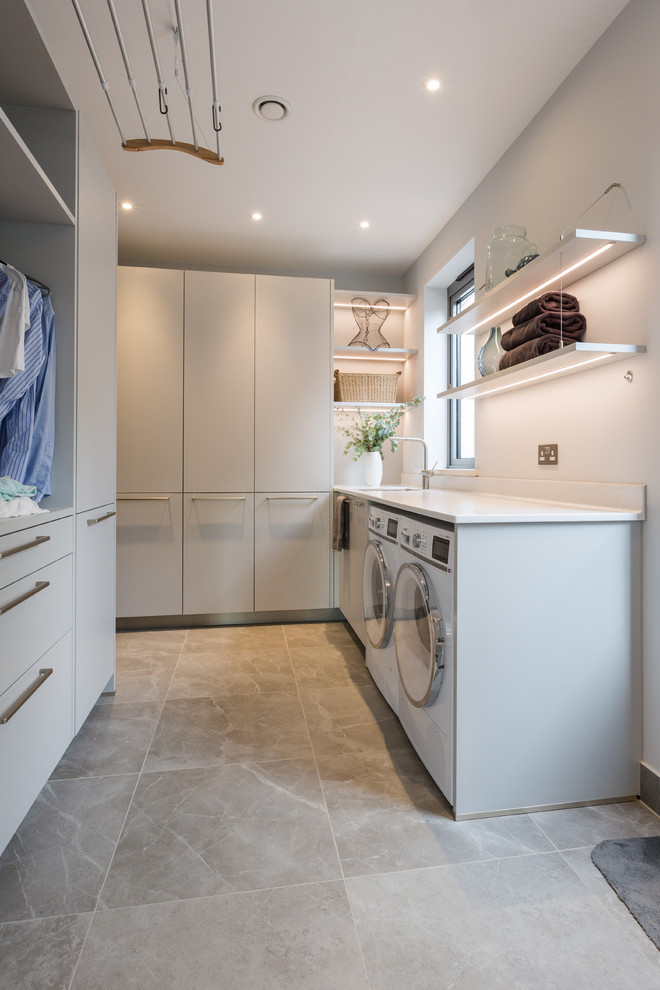 Medium sized modern u-shaped separated utility room in Gloucestershire with a built-in sink, flat-panel cabinets, composite countertops, metallic splashback, glass sheet splashback, ceramic flooring, brown floors, grey cabinets, white walls, a side by side washer and dryer and white worktops.
