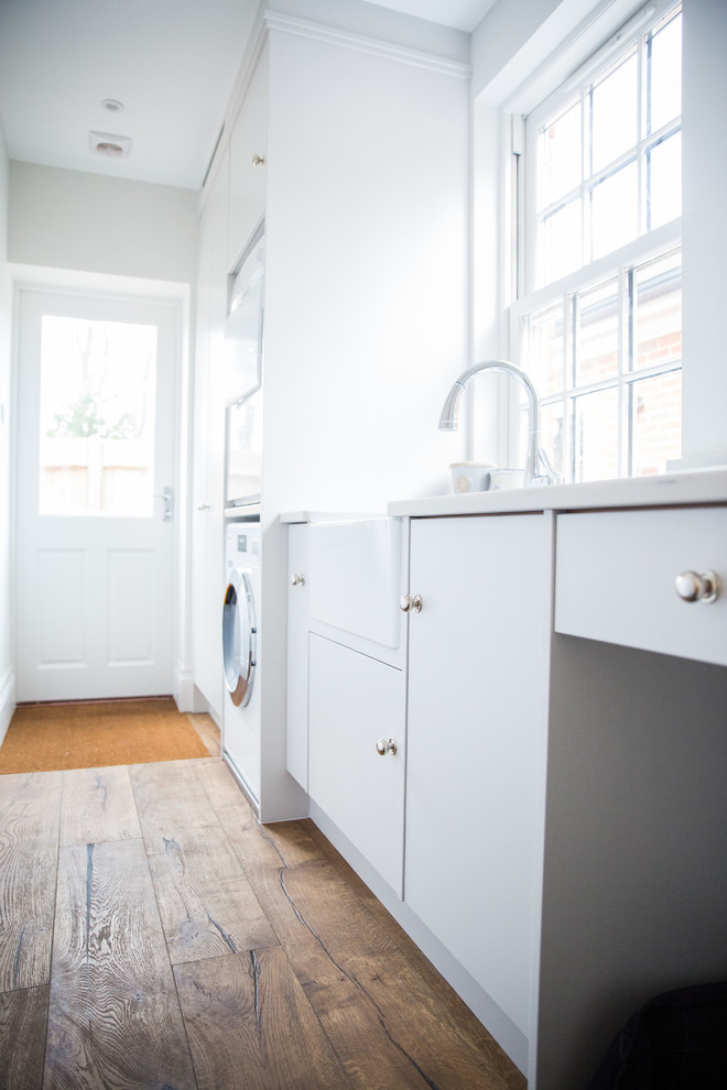 Medium sized modern galley utility room in Berkshire with a belfast sink, flat-panel cabinets, white cabinets, composite countertops, white walls, dark hardwood flooring, a stacked washer and dryer, brown floors and white worktops.