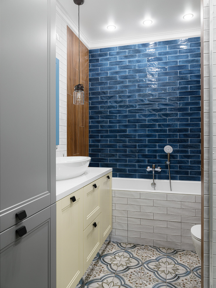 На фото: ванная комната в современном стиле с желтыми фасадами, ванной в нише, душем над ванной, синей плиткой, настольной раковиной, разноцветным полом и белой столешницей с