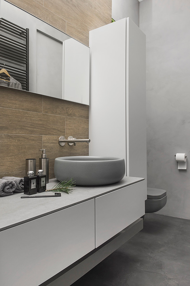 Exempel på ett industriellt badrum, med släta luckor, vita skåp, en vägghängd toalettstol, brun kakel, grå väggar och ett fristående handfat