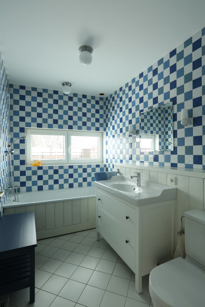 Modelo de cuarto de baño principal escandinavo con armarios con paneles lisos, puertas de armario blancas, bañera empotrada, sanitario de dos piezas, baldosas y/o azulejos azules, baldosas y/o azulejos blancos y lavabo integrado