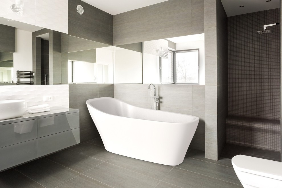 Idée de décoration pour une grande salle de bain design avec une baignoire indépendante, WC suspendus, un carrelage gris, des dalles de pierre, un mur beige, tomettes au sol, une vasque et un plan de toilette en surface solide.
