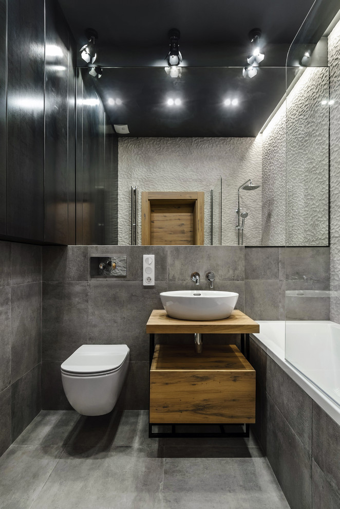 Diseño de cuarto de baño urbano, de tamaño medio, con armarios con paneles lisos, puertas de armario de madera oscura, combinación de ducha y bañera, sanitario de pared, baldosas y/o azulejos grises, baldosas y/o azulejos de porcelana, suelo de baldosas de porcelana, encimera de madera, suelo gris, bañera empotrada, lavabo sobreencimera, ducha abierta, encimeras marrones, paredes grises y aseo y ducha