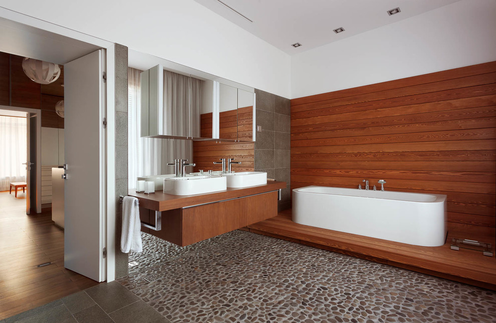Modelo de cuarto de baño contemporáneo con armarios con paneles lisos, bañera exenta, suelo de baldosas tipo guijarro, suelo de baldosas tipo guijarro, encimera de madera y encimeras marrones