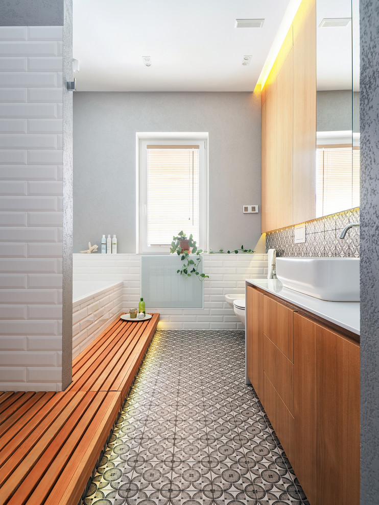 Modernes Badezimmer mit flächenbündigen Schrankfronten, hellbraunen Holzschränken, Einbaubadewanne, Bidet, farbigen Fliesen, grauer Wandfarbe, Aufsatzwaschbecken, buntem Boden und weißer Waschtischplatte in Sonstige