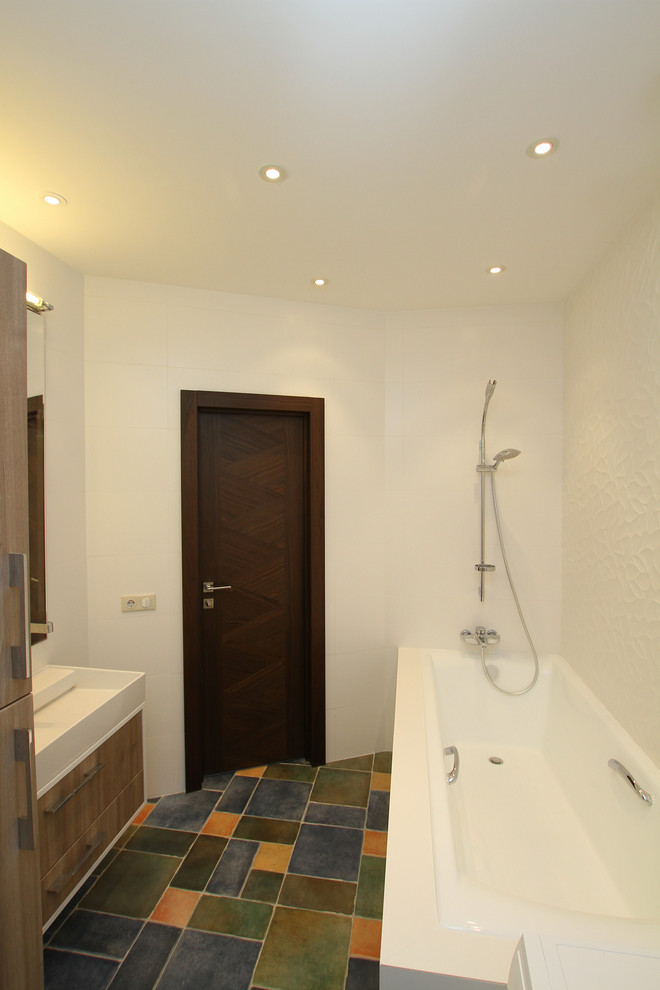 This is an example of a medium sized contemporary ensuite bathroom in Moscow with flat-panel cabinets, medium wood cabinets, a submerged bath, a shower/bath combination, a wall mounted toilet, white tiles, ceramic tiles, white walls, ceramic flooring, a built-in sink, multi-coloured floors and white worktops.