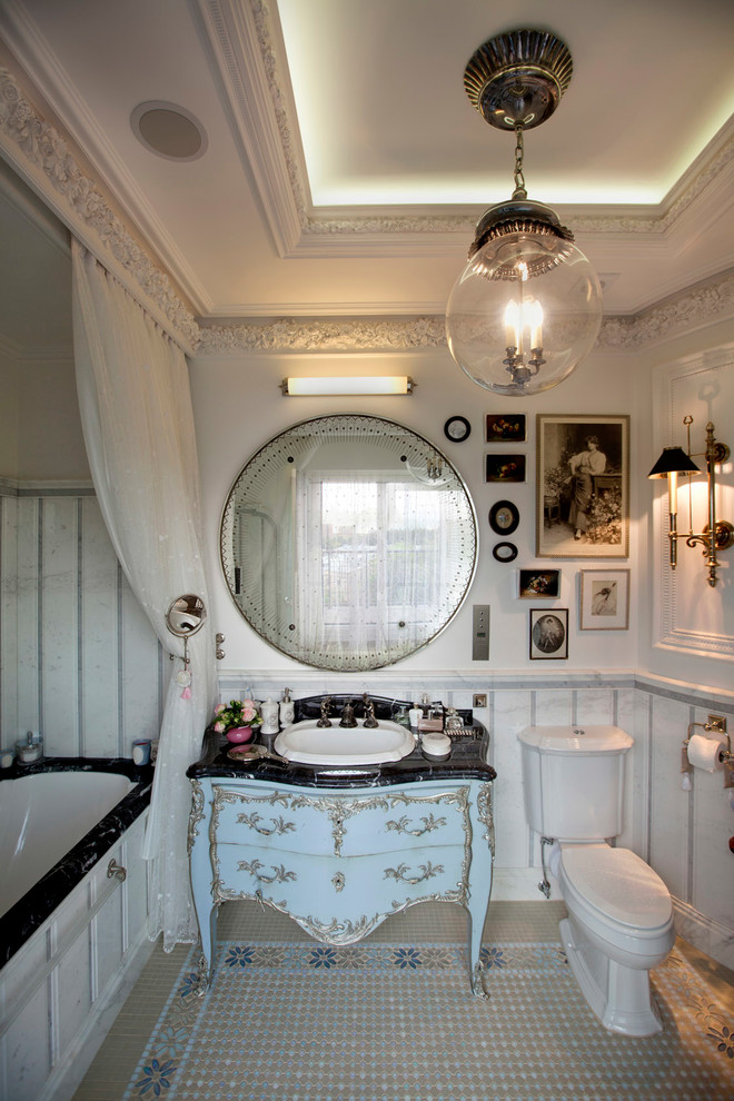 На фото: главная ванная комната в стиле неоклассика (современная классика) с синими фасадами, полновстраиваемой ванной, белыми стенами, накладной раковиной и черной столешницей с