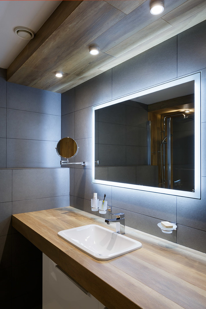 На фото: главная ванная комната в современном стиле с плоскими фасадами, белыми фасадами, серой плиткой, керамогранитной плиткой, накладной раковиной, столешницей из дерева, коричневой столешницей и зеркалом с подсветкой