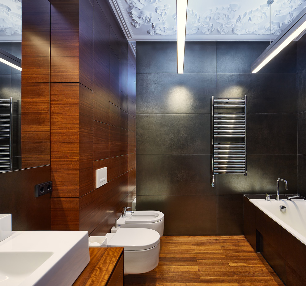 Aménagement d'une salle de bain principale contemporaine de taille moyenne avec un placard à porte plane, des portes de placard marrons, une baignoire encastrée, WC suspendus, un carrelage gris, carrelage en métal, un mur gris, un sol en bois brun, une vasque et un plan de toilette en bois.