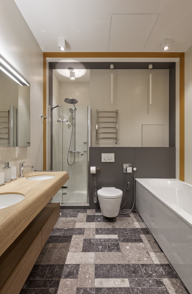 Idéer för att renovera ett mellanstort funkis beige beige en-suite badrum, med grå kakel, stenhäll, släta luckor, skåp i ljust trä, ett badkar i en alkov, en hörndusch, en vägghängd toalettstol, vita väggar, ett undermonterad handfat, träbänkskiva, grått golv och dusch med gångjärnsdörr