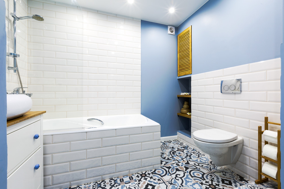 Aménagement d'une petite salle de bain scandinave avec un placard à porte plane, des portes de placard blanches, un combiné douche/baignoire, WC suspendus, des carreaux de céramique, un sol en carrelage de céramique, un plan de toilette en bois, un sol multicolore, un carrelage blanc, un mur bleu, une vasque et un plan de toilette marron.