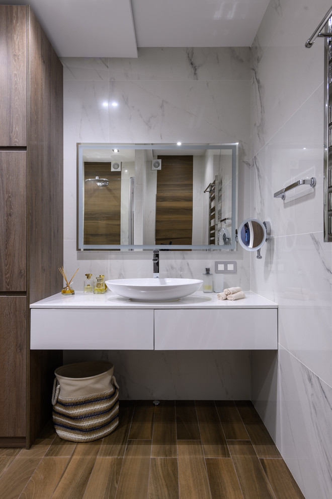 Пример оригинального дизайна: ванная комната среднего размера в стиле неоклассика (современная классика) с плоскими фасадами, белыми фасадами, душем в нише, инсталляцией, белой плиткой, керамогранитной плиткой, белыми стенами, полом из керамогранита, душевой кабиной, настольной раковиной, столешницей из искусственного камня, коричневым полом, душем с распашными дверями и белой столешницей