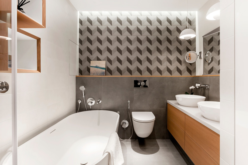 Inredning av ett modernt en-suite badrum, med släta luckor, skåp i mellenmörkt trä, ett fristående badkar, en vägghängd toalettstol, vit kakel, grå kakel och ett fristående handfat