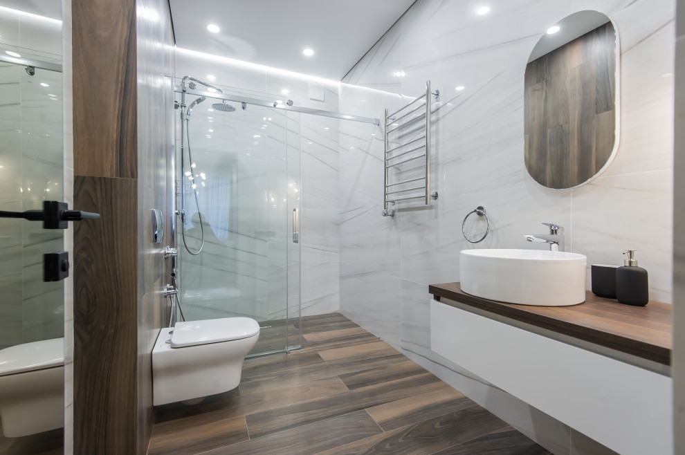 Idéer för ett stort modernt brun badrum med dusch, med släta luckor, vita skåp, en kantlös dusch, en vägghängd toalettstol, vit kakel, porslinskakel, klinkergolv i porslin, ett fristående handfat, träbänkskiva, brunt golv och dusch med skjutdörr