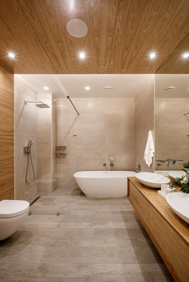 Bild på ett stort funkis beige beige en-suite badrum, med släta luckor, beige skåp, ett fristående badkar, våtrum, en vägghängd toalettstol, beige kakel, porslinskakel, bruna väggar, ett fristående handfat, träbänkskiva, grått golv och med dusch som är öppen