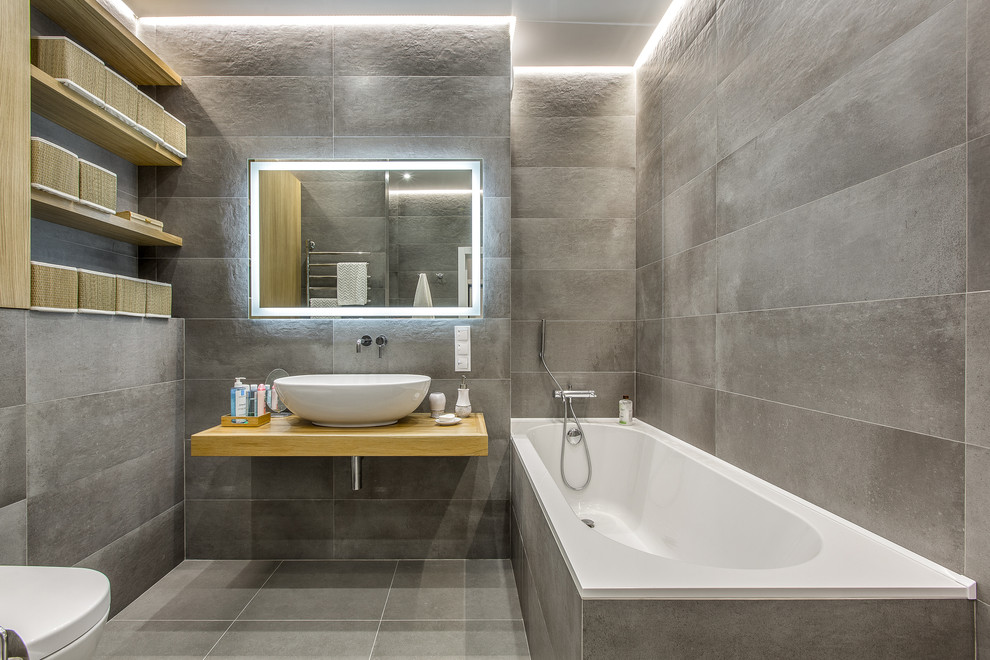 На фото: главная ванная комната: освещение в современном стиле с открытыми фасадами, душем над ванной, раздельным унитазом, серой плиткой, настольной раковиной, столешницей из дерева и бежевой столешницей с