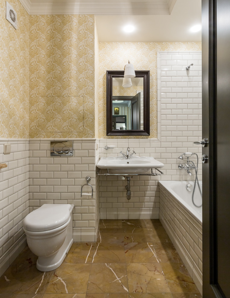 На фото: главная ванная комната в стиле неоклассика (современная классика) с ванной в нише, душем над ванной, раздельным унитазом, белой плиткой, плиткой кабанчик, желтыми стенами, подвесной раковиной и коричневым полом