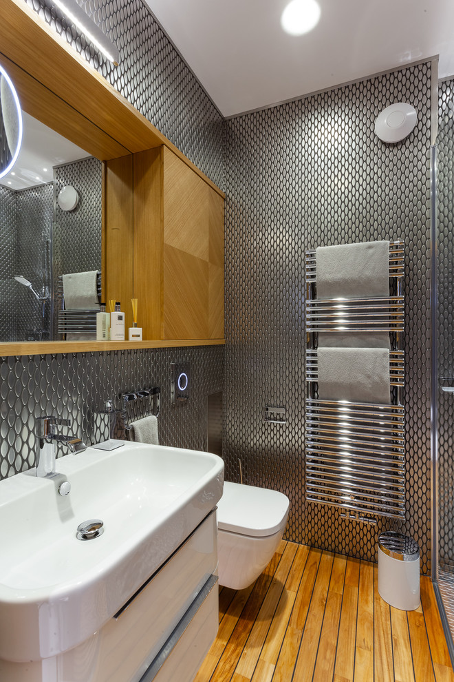 Cette photo montre une salle d'eau tendance avec un placard à porte plane, des portes de placard blanches, WC suspendus, carrelage en métal, un sol en bois brun et un lavabo suspendu.