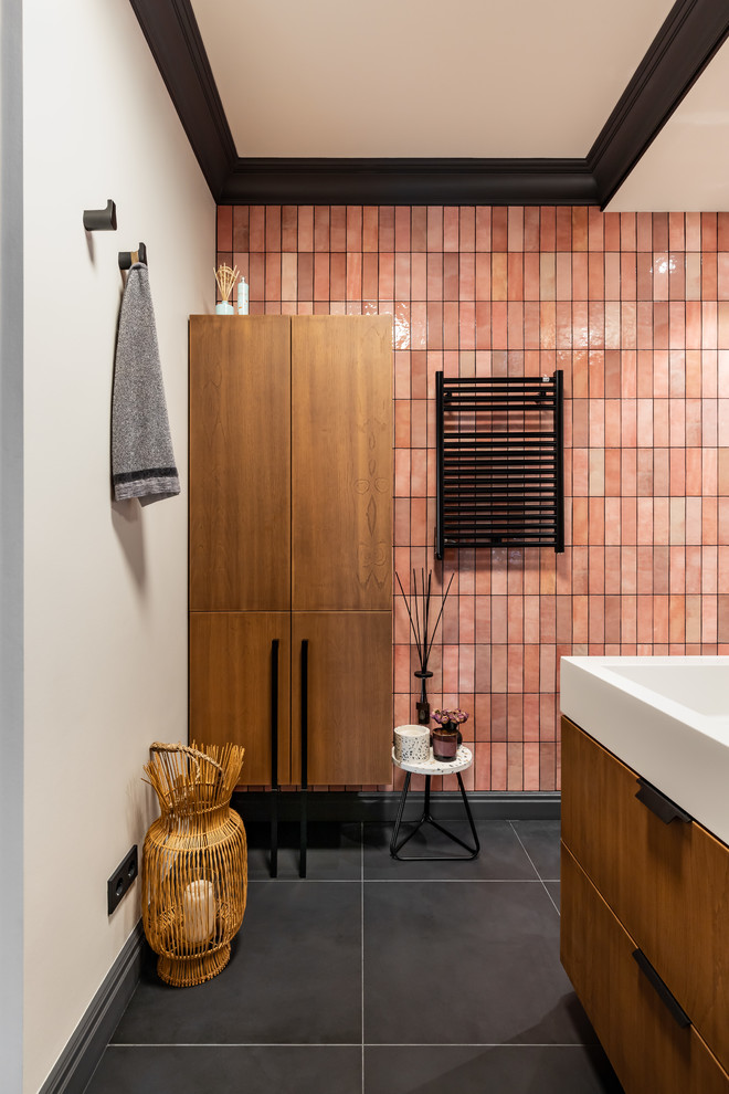 Inredning av ett badrum, med släta luckor, skåp i mellenmörkt trä, ett undermonterat badkar, en vägghängd toalettstol, rosa kakel, keramikplattor, rosa väggar, klinkergolv i porslin, ett väggmonterat handfat och svart golv