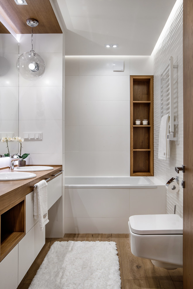 Idéer för små funkis en-suite badrum, med släta luckor, vita skåp, ett badkar i en alkov, en dusch/badkar-kombination, en vägghängd toalettstol, vit kakel, vita väggar, klinkergolv i porslin, träbänkskiva, ett nedsänkt handfat och brunt golv