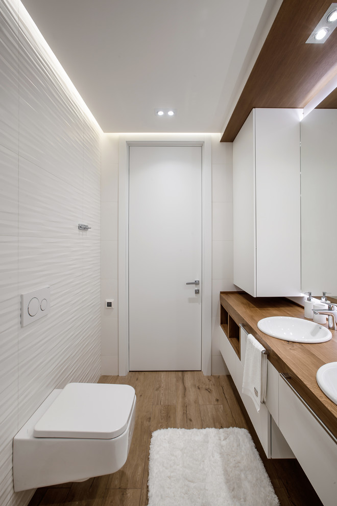 Inredning av ett modernt litet en-suite badrum, med släta luckor, vita skåp, ett badkar i en alkov, en dusch/badkar-kombination, en vägghängd toalettstol, vit kakel, stickkakel, vita väggar, klinkergolv i porslin, ett undermonterad handfat och träbänkskiva