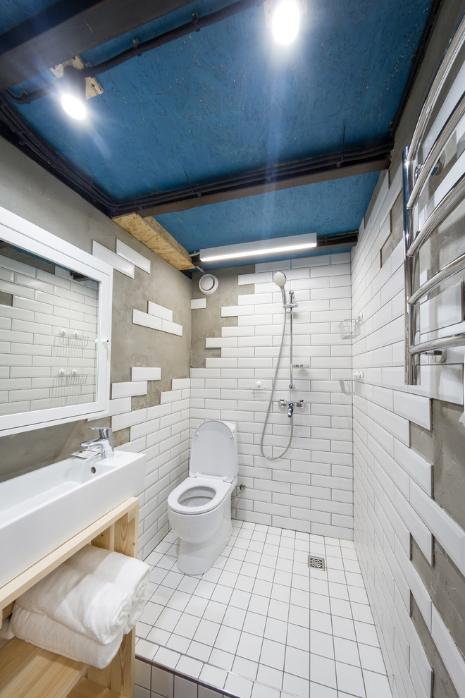 Inredning av ett industriellt litet badrum med dusch, med öppna hyllor, skåp i ljust trä, en kantlös dusch, en toalettstol med separat cisternkåpa, vit kakel, grå väggar, ett fristående handfat och med dusch som är öppen