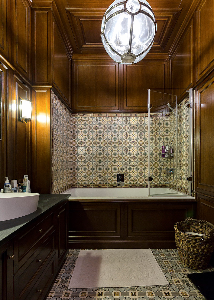 На фото: ванная комната в классическом стиле с темными деревянными фасадами, разноцветной плиткой, коричневыми стенами, полом из керамической плитки, настольной раковиной, ванной в нише и душем над ванной