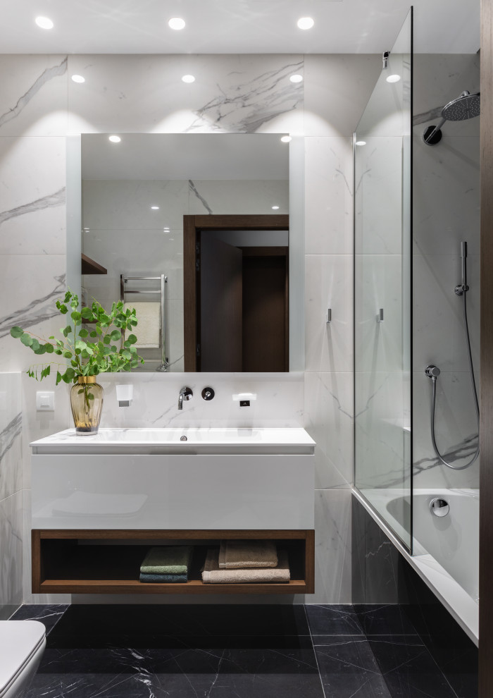 На фото: ванная комната в современном стиле с плоскими фасадами, белыми фасадами, ванной в нише, душем над ванной, белой плиткой, монолитной раковиной, черным полом, открытым душем, белой столешницей, тумбой под одну раковину и подвесной тумбой