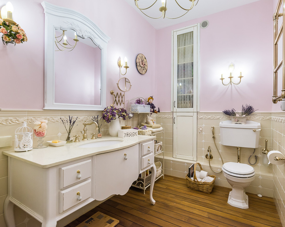 Aménagement d'une salle de bain classique avec des portes de placard blanches, WC séparés, un carrelage beige, un mur rose, un lavabo encastré, un sol en bois brun et un placard avec porte à panneau surélevé.