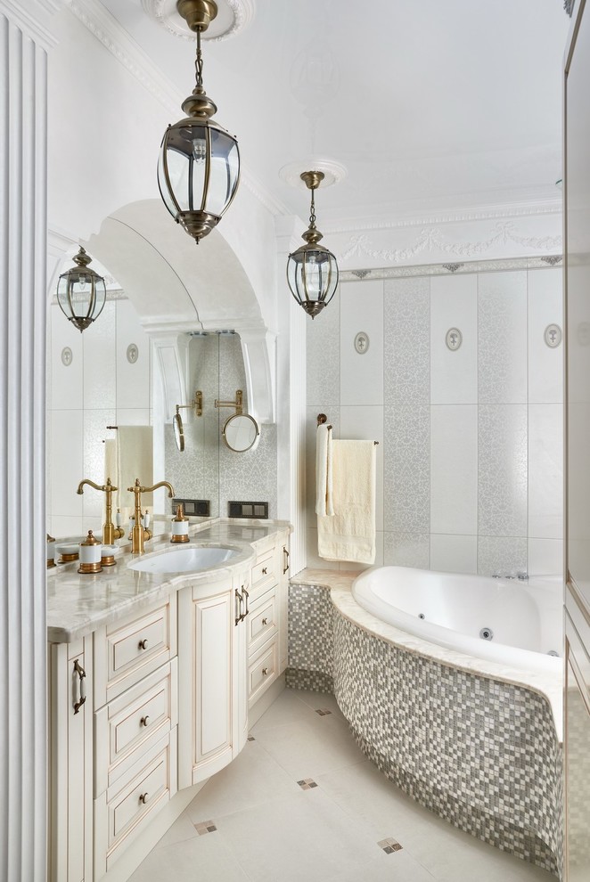 Cette photo montre une salle de bain victorienne avec un placard avec porte à panneau surélevé, des portes de placard beiges, un mur blanc, un lavabo encastré, un bain bouillonnant, un carrelage blanc et un carrelage gris.