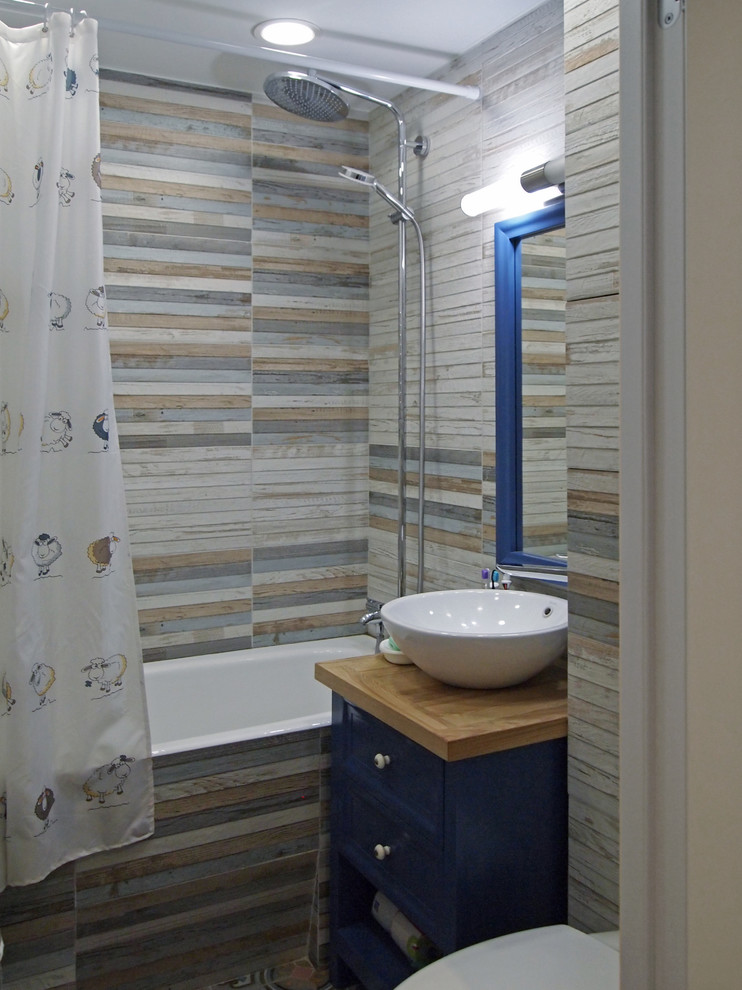 Foto på ett litet skandinaviskt en-suite badrum, med luckor med infälld panel, blå skåp, en vägghängd toalettstol, flerfärgad kakel, porslinskakel, träbänkskiva, ett badkar i en alkov och ett fristående handfat