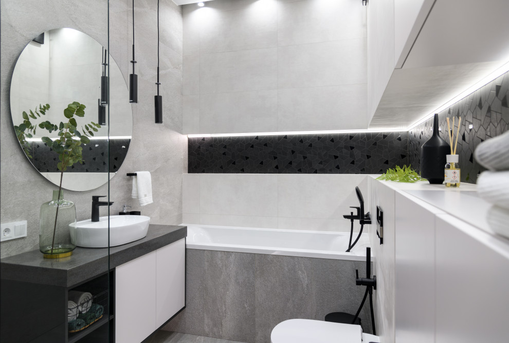 Пример оригинального дизайна: большая главная ванная комната в современном стиле с плоскими фасадами, белыми фасадами, полновстраиваемой ванной, угловым душем, инсталляцией, серой плиткой, керамогранитной плиткой, серыми стенами, полом из керамогранита, накладной раковиной, столешницей из искусственного кварца, серым полом, душем с раздвижными дверями, серой столешницей, тумбой под одну раковину и подвесной тумбой