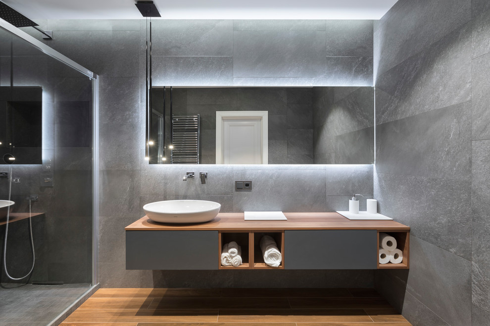 Modelo de cuarto de baño actual con puertas de armario grises, ducha empotrada, baldosas y/o azulejos grises, paredes grises, aseo y ducha, lavabo sobreencimera, encimera de madera y suelo marrón