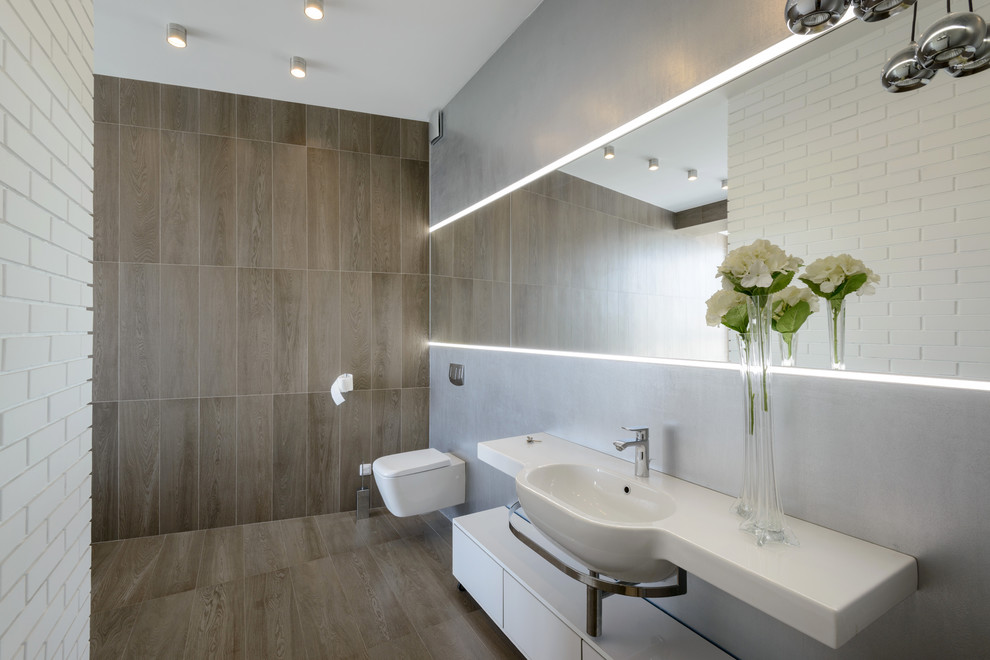 Imagen de cuarto de baño contemporáneo con sanitario de pared, paredes grises, suelo de madera en tonos medios, lavabo integrado y espejo con luz