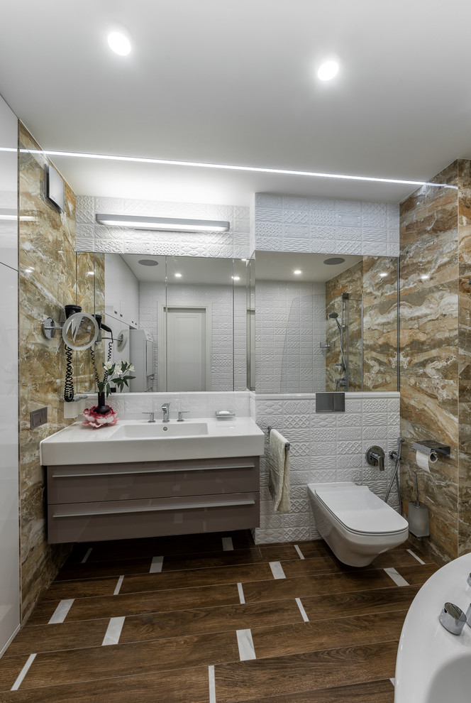 Exemple d'une grande salle de bain principale tendance avec un placard à porte plane, des portes de placard marrons, WC suspendus, un carrelage beige, un carrelage blanc, des carreaux de porcelaine, un mur beige, un sol en carrelage de porcelaine, un plan de toilette en surface solide, un combiné douche/baignoire et un lavabo intégré.