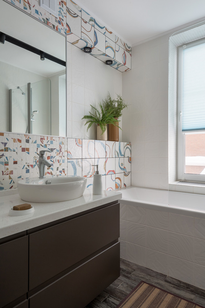 Cette image montre une salle de bain principale design avec un placard à porte plane, des portes de placard marrons, une baignoire en alcôve, un carrelage blanc, un carrelage multicolore, une vasque, un sol gris et un plan de toilette blanc.