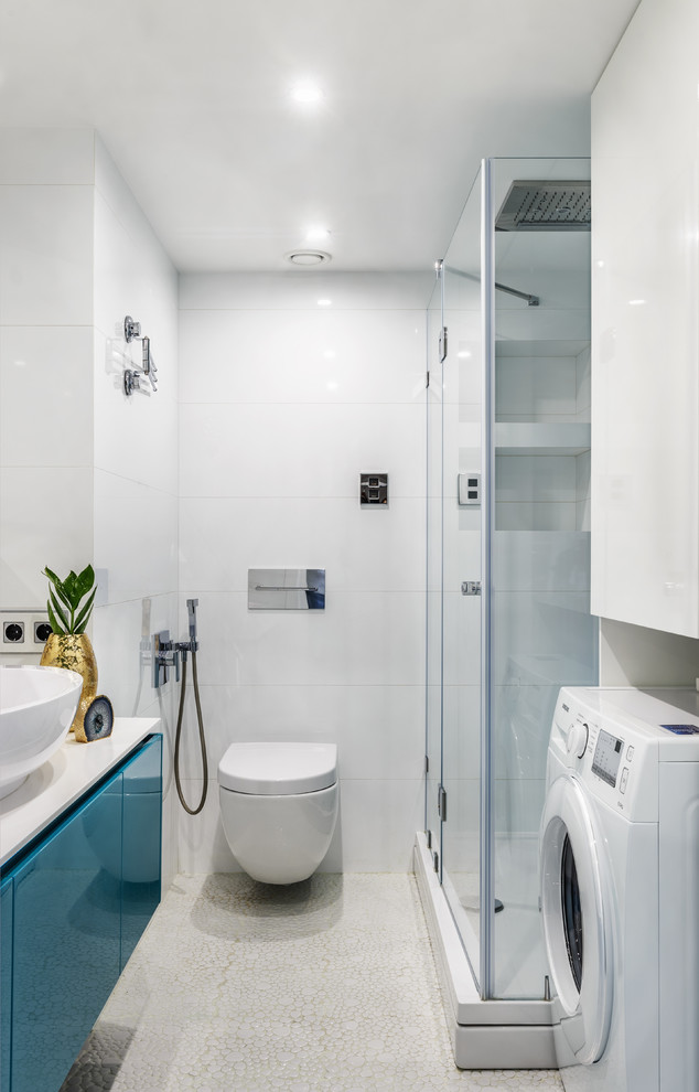 Cette image montre une salle d'eau design avec un placard à porte plane, des portes de placard bleues, une douche d'angle, WC suspendus, un carrelage blanc, une vasque, une cabine de douche à porte battante, un sol en galet et un sol blanc.