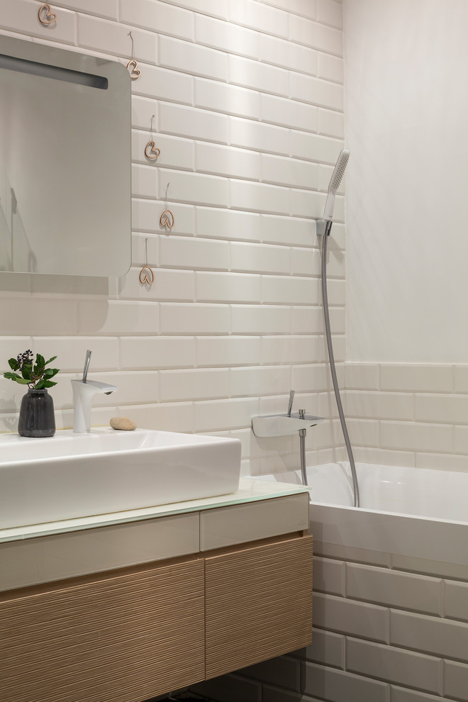 На фото: главная ванная комната в современном стиле с плоскими фасадами, светлыми деревянными фасадами, ванной в нише, душем над ванной, плиткой кабанчик, настольной раковиной и стеклянной столешницей с