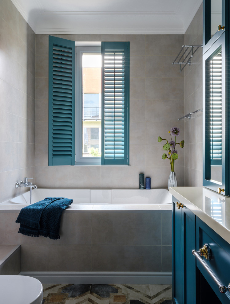 Foto på ett mellanstort vintage beige en-suite badrum, med grå kakel, porslinskakel, klinkergolv i porslin, bänkskiva i akrylsten, flerfärgat golv, ett badkar i en alkov, skåp i shakerstil, blå skåp och grå väggar