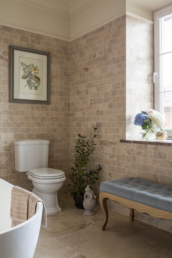 Foto på ett mellanstort vintage badrum, med ett fristående badkar, en toalettstol med separat cisternkåpa, beige kakel, marmorkakel och beiget golv